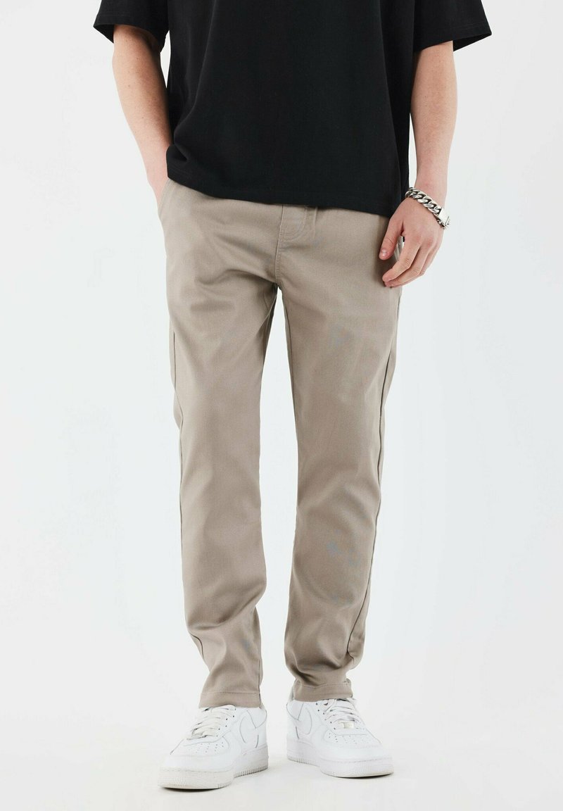 Chinos beiges ajustés, en tissu mélangé de coton. Comprend des poches latérales et une texture lisse, associés à des baskets blanches.