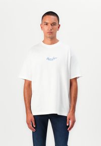 Weißes Baumwoll-T-Shirt mit lockerer Passform, rundem Ausschnitt und kurzen Ärmeln. Mit blauem "Guess Jeans"-Logo auf der Brust. Getragen mit dunklen Jeans.