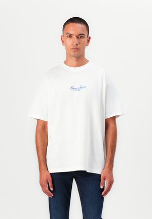 Weißes Baumwoll-T-Shirt mit lockerer Passform, rundem Ausschnitt und kurzen Ärmeln. Mit blauem "Guess Jeans"-Logo auf der Brust. Getragen mit dunklen Jeans.