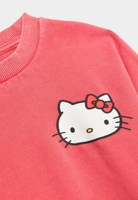 Sudadera de forro polar en coral, con un gráfico de Hello Kitty en blanco y negro y un lazo rojo. Cuello acanalado y textura suave.