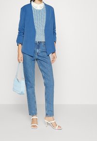 Modrý blejzr, světle modrý pletený svetr s copánkovým vzorem, vybledlé modré džíny, bílé sandály na podpatku s pásky a světle modrá prošívaná kabelka.