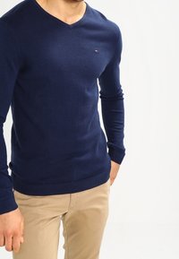 Man som bär en marinblå V-ringad tröja och beige byxor, stående mot en enkel vit bakgrund.