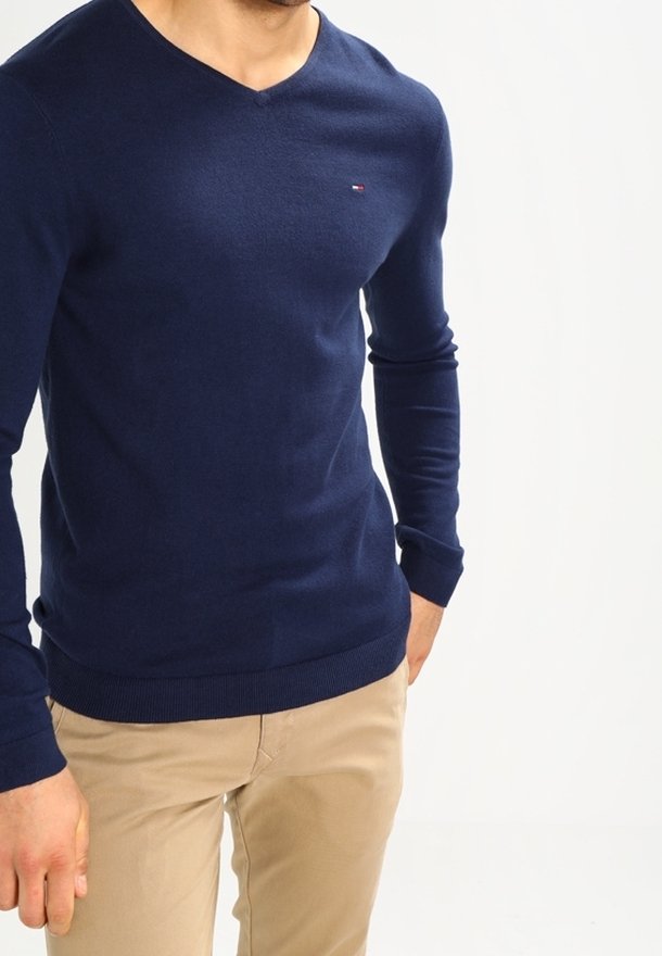 Man som bär en marinblå V-ringad tröja och beige byxor, stående mot en enkel vit bakgrund.