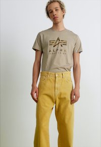 Bežinis marškinėliai su "ALPHA INDUSTRIES" logotipu, kuriame yra maskuotė, derinami su aukštu liemeniu geltonais džinsais su klasikiniu siluetu.