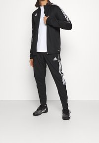 Man som bär en svart Adidas träningsjacka, vit t-shirt, svarta Adidas träningsbyxor och svarta skor med vita fläckar, stående på ett vitt golv.