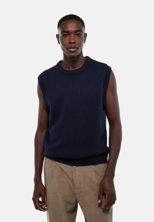 Pull en tricot sans manches bleu marine avec un col et un ourlet côtelés, fabriqué à partir d'un tissu texturé. Porté sur un pantalon en velours côtelé beige.