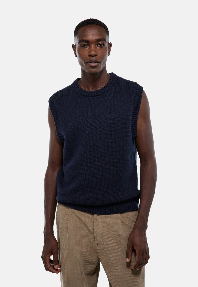 Pull en tricot sans manches bleu marine avec un col et un ourlet côtelés, fabriqué à partir d'un tissu texturé. Porté sur un pantalon en velours côtelé beige.