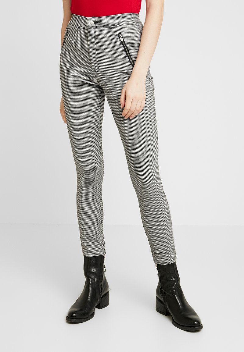 Pantalon slim noir et blanc à carreaux avec poche zippée à l'avant, porté avec des bottines noires et un haut rouge.