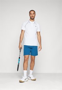 Bílá polo triko s černými detaily, modré sportovní šortky a bílé tenisky. Model drží tenisovou raketu a stojí na jednobarevném pozadí.
