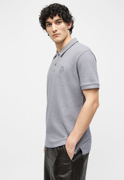 Νεαρός με σγουρά μαλλιά φοράει ανοιχτό γκρι κοντομάνικο πόλο μπλουζάκι και μαύρο υφασμάτινο παντελόνι, με το ένα χέρι στην τσέπη του.