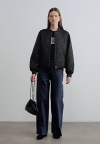 Μαύρο καπιτονέ bomber μπουφάν με ριμπ γιακά, συνδυασμένο με σκούρο παντελόνι φαρδιάς γραμμής. Το μοντέλο κρατά μικρή μαύρη τσάντα τύπου bucket με μινιμαλιστικό σχέδιο.