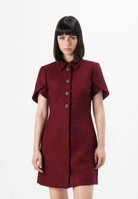Donna che indossa un vestito aderente a maniche corte color bordeaux con bottoni e colletto, in piedi contro uno sfondo bianco uniforme.