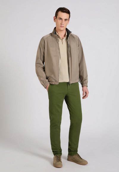 Man staat met handen in de zakken, draagt een beige jas, lichtbeige polo, olijfgroene broek en beige sneakers tegen een effen achtergrond.