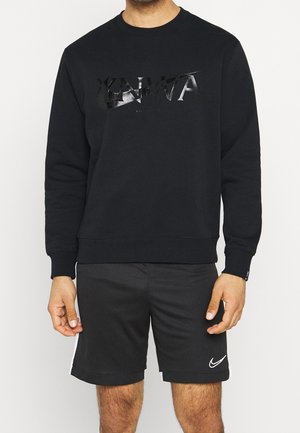 Mężczyzna w czarnej bluzie z długim rękawem z napisem „YNWA” oraz czarnych szortach Nike z białym logo „swoosh” na lewej nodze.