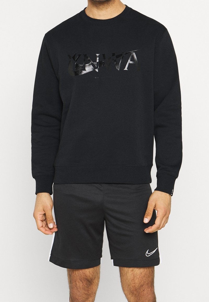 Homme portant un sweat à manches longues noir avec l’inscription « YNWA » et un short noir Nike avec le logo swoosh blanc sur la jambe gauche.