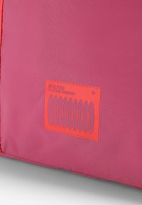 Bolso de tela rosa que presenta una etiqueta rectangular de color naranja vibrante con texto gris y patrones elípticos. Textura suave y bordes cosidos.