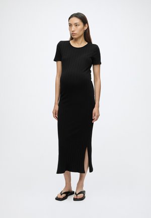 Femme enceinte debout, portant une robe midi noire côtelée à manches courtes avec une fente latérale et des sandales plates noires sur fond blanc.