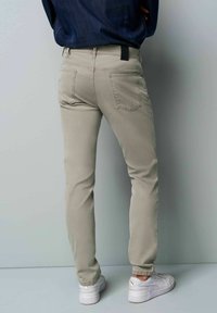 Beige Slim-Fit-Hosen mit Gesäßtaschen, versehen mit einem schwarzen Markenetikett am Bund. Kombiniert mit weißen Sneakers. Glatte Textur.