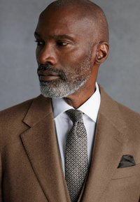Brown wool suit jacket with notch lapels, white shirt, patterned tie, and dark pocket square. Close-up view focuses on fabric textures and details.