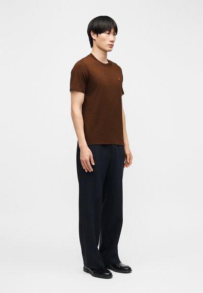 Jeune homme debout, portant un t-shirt marron à manches courtes, un pantalon large sombre et des chaussures habillées noires, sur un fond clair uni.