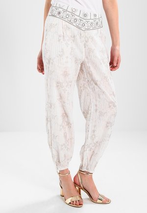 Kobieta nosząca białe, luźne spodnie z wzorem, z haftowanym pasem i złotymi sandałami na wysokim obcasie na białym tle.