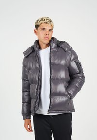 Geïsoleerde jas in metallic grijs met een rits aan de voorkant, afneembare capuchon en elastische manchetten. Voorzien van zijzakken en een ritszak op de arm.
