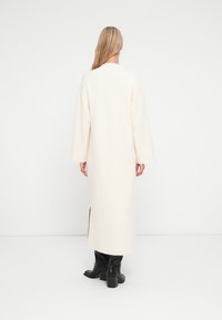 Robe longue de couleur crème avec une coupe ample, un col rond et une fente sur le côté, associée à des bottes à talons noires. Texture douce, design minimal.