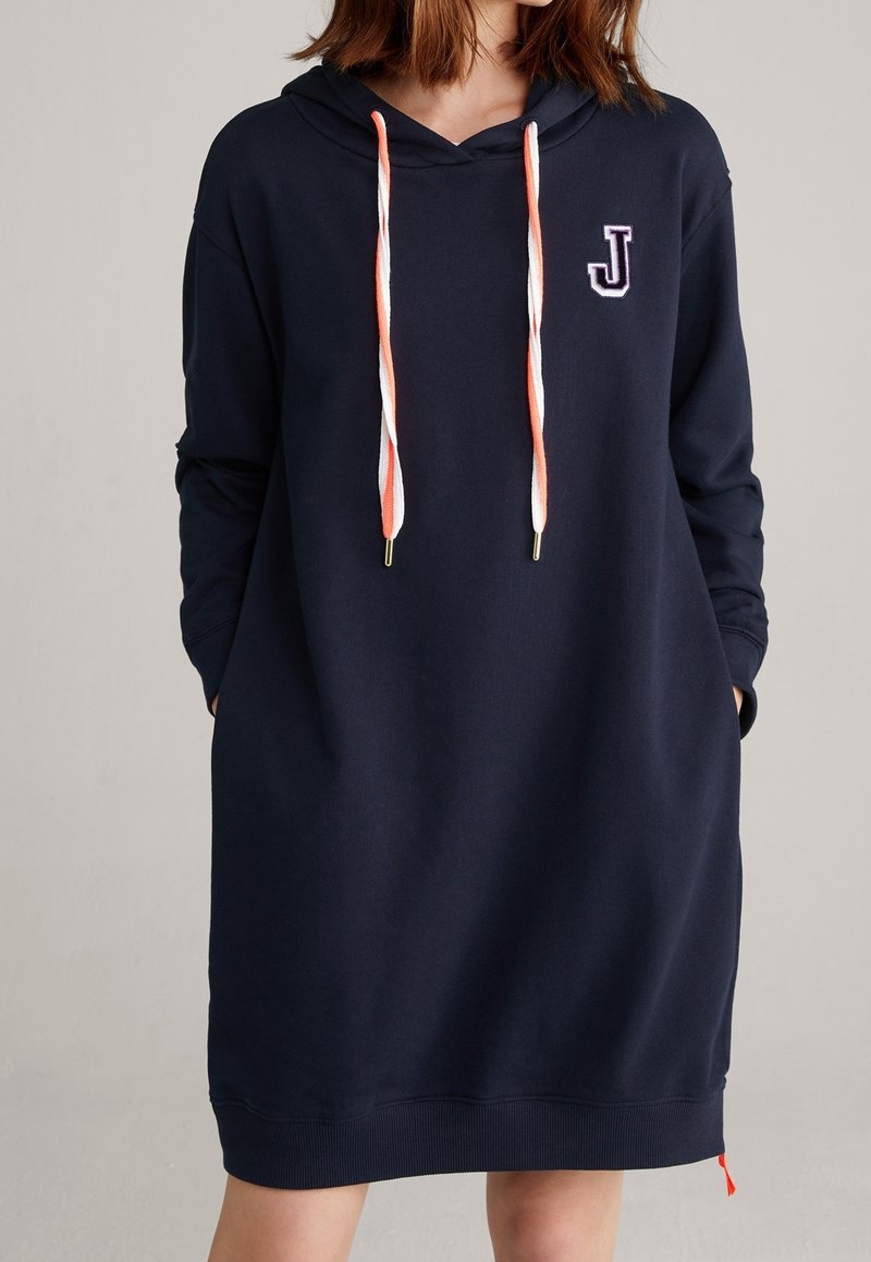 Femme portant une robe longue à capuche bleu marine avec des cordons orange et blancs et un écusson blanc « J » sur la poitrine gauche, mains dans les poches.