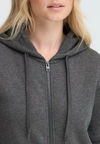 Felpa grigia con zip e cappuccio regolabile, caratterizzata da una superficie liscia e una cerniera in metallo, vista dalla parte superiore del torace.