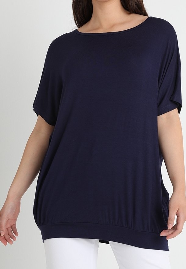Femme portant un haut ample bleu marine à manches courtes avec un large col et un pantalon blanc sur un fond uni.