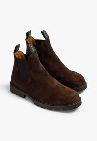 Las botas Chelsea de ante marrón presentan paneles laterales elásticos, lengüetas de tiro y una suela de goma negra con un patrón texturizado para mayor agarre.