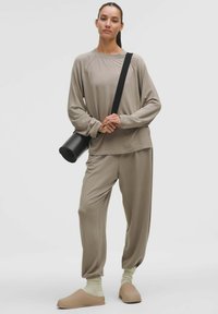 Set de îmbrăcăminte de casă bej, format dintr-un top lejer cu mânecă lungă și pantaloni tapered, asortați cu papuci ușori și șosete verzuie. Geantă mată neagră.