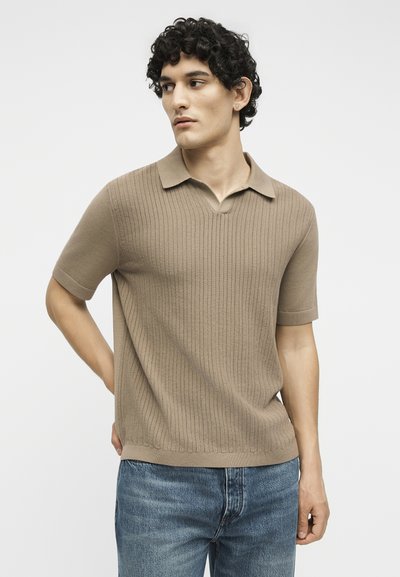 Νεαρός άνδρας με σγουρά μαλλιά που φοράει μια μπεζ κοντομάνικη πλεκτή πόλο μπλούζα και μπλε τζιν, κοιτάζοντας προς τα αριστερά με φόντο ένα απλό χρώμα.