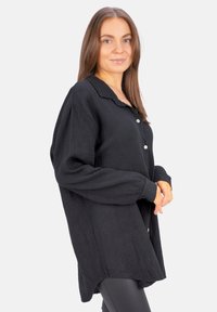 Black button-up shirt made of textured fabric, featuring a collar, long sleeves, and a slightly oversized fit with a curved hemline.