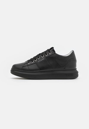Baskets basses en cuir noir avec lacets, œillets argentés et marquage "GUESS" sur le côté et la semelle, présentées en vue de profil côté droit.