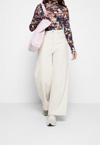 Weite cremefarbene Hosen mit hoher Taille, kombiniert mit einem floralem Langarmoberteil in dunklen Farben und einer sanften rosa Tote-Bag.