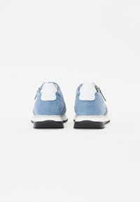 Baskets en denim bleu clair avec accents blancs, vue arrière. Présentent une texture lisse, une semelle noire contrastante et un détail de fermeture éclair.