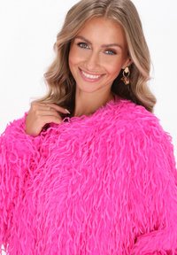 Fluffy roze sweater met lange strengen, met een losse pasvorm. Model draagt een paar grote oorbellen. De gladde achtergrond accentueert de textuur van het kledingstuk.