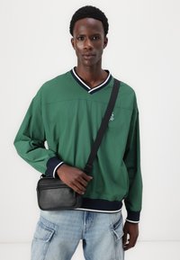 Cămașă verde ușoară cu mânecă lungă, cu guler în V și tiv bleumarin; purtată împreună cu o geantă de umăr rectangulară neagră și pantaloni din denim de culoare bleu deschis.