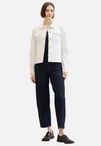 White denim jacket with two chest pockets, button closure. Worn over a black top and paired with dark blue, loose-fitting jeans and black shoes.