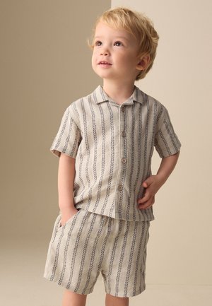 Ung blondt barn iført matchende beige kortærmet skjorte og shorts med mørke lodrette bladmønstre, kigger op med hænderne i lommerne.