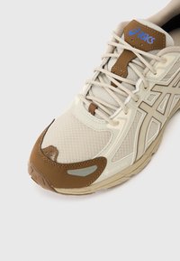 Beidzītā krāsā un brūnā Asics skriešanas apavi ar sietu audumu, sintētiskajām virsmām, auklām un Gore-Tex marķējumu uz baltā fona.