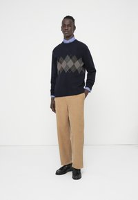 Vīrietis stāv baltā fonā, valkājot bēšas kordurojas bikses, melnu džemperi ar pelēku argaila rakstu, zilu rūtainu kreklu un melnas loafer apavus.