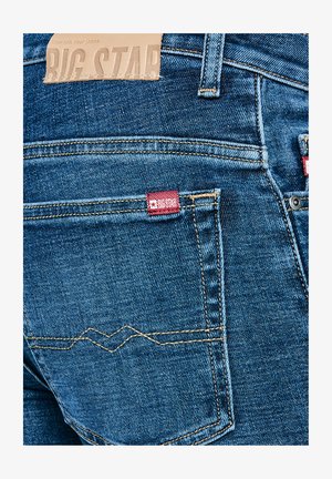 Blå denimjeans med baglomme, der viser gul syning, rød Big Star-mærkat og beige læderlabel ved taljen.