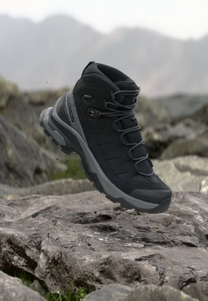Zwarte Salomon wandelschoen met Gore-Tex zool zwevend boven rotsachtig terrein met een vervaagde bergachtergrond.