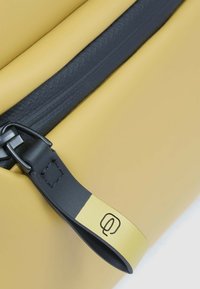 Gelbe Tasche mit strukturierter Oberfläche, schwarzem Reißverschluss und einem schwarzen Riemen mit gelbem Akzent und eingeprägtem Logodetail.