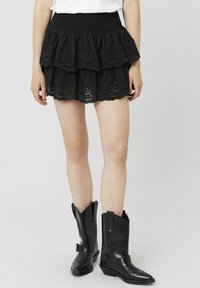 Jupe mini noire à volants en tissu œillet, avec un ourlet festonné et une taille smockée. Associée à des bottes de cowboy noires à bout pointu.