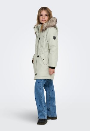 Svijetlozeleno parka s umjetnim krznom na kapuljači, vezicama u struku, višestrukim gumbima i crnom logotip oznakom. Nošeno s širokim plavim trapericama i crnim čizmama.