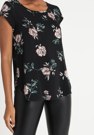 Zwarte bloemenprint korte mouwen blouse met roze en groene bloemen, gedragen met zwarte leren broek, getoond op een staande persoon van hals tot dij.