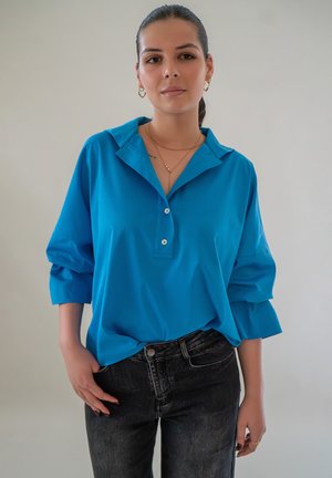 Blauwe knopshirt met een brede kraag en driekwart mouwen, gecombineerd met donkere spijkerbroek. Zachte stof met een gladde textuur.
